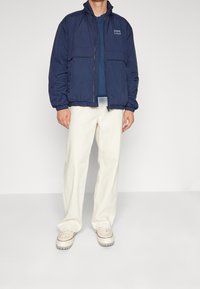 Marinblå lättviktsjacka med hög krage, frontdragkedja, elastiska ärmar och två framfickor, ihop med krämfärgade byxor.