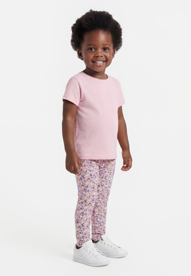 Uśmiechnięta dziewczynka w wieku niemowlęcym, ubrana w jasno różową krótką koszulkę, kwiatowe legginsy i białe tenisówki, stojąca na jednolitym tle.