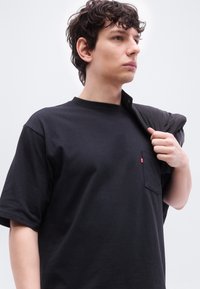 Νεαρός άνδρας με κοντά σγουρά μαλλιά, που φοράει μαύρο μπλουζάκι, κρατώντας ένα μαύρο σακάκι στον ώμο του, κοιτάζοντας προς τα δεξιά.