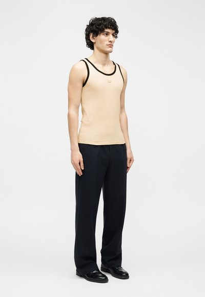 Jeune homme debout, portant un débardeur beige avec bordure noire, un pantalon large noir et des chaussures habillées noires sur un fond clair uni.