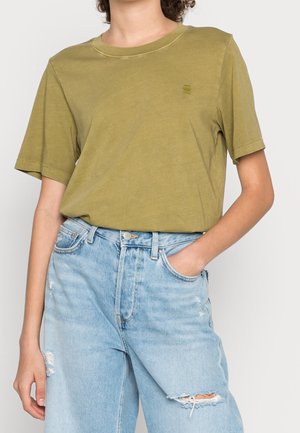 Person iført en olivengrøn T-shirt med korte ærmer, stoppet ned i lyseblå distressed denimjeans, med den ene hånd i lommen.