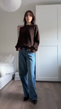 Brauner strukturierter Pullover, locker sitzende hellblaue Jeans, spitze schwarze Absätze. Steht in einem Raum mit hellem Holzboden und weißen Möbeln.