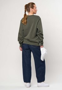 Femeie cu păr împletit, purtând hanorac verde, blugi albastru închis, adidași albi, ținând o șapcă albă, stând în fața unui fundal simplu.