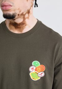 Oliven grøn bomulds T-shirt med et sæt farverige Adidas logo klistermærker i forskellige former, placeret på venstre brystområde.