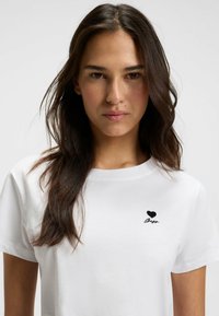 White cotton t-shirt featuring a black heart logo and script text on the left chest area; round neckline, short sleeves, smooth texture.