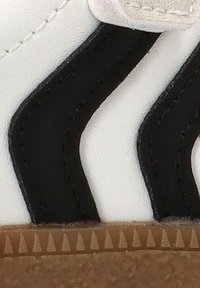 Zapatilla blanca con bandas en zigzag negras, suela de goma texturizada con pequeños patrones triangulares y detalles de costura en contraste.