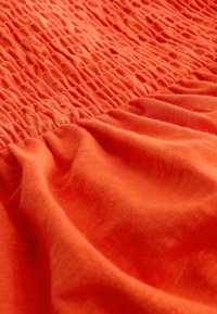 Gros plan d’un tissu orange vif avec des fronces et une couture smockée sur la partie supérieure.