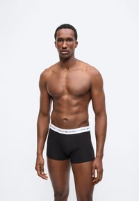 Μυώδης άνδρας με μαύρα εσώρουχα Tommy Hilfiger στέκεται μπροστά σε απλό λευκό φόντο, κοιτάζοντας μπροστά με ουδέτερη έκφραση.