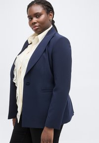 Αυτοπεποίθητη γυναίκα με πλεξούδες, που φοράει μπλε σακάκι, κρεμ μπλούζα με βολάν και χρυσά σκουλαρίκια, στέκεται μπροστά σε απλό φωτεινό φόντο.