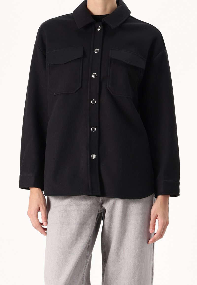 Veste noire à boutons avec col et deux poches poitrine portée avec un jean gris clair sur une personne debout.