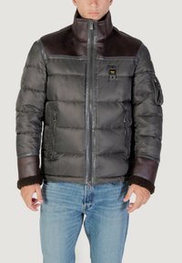 Uomo che indossa un giubbotto imbottito nero con dettagli in pelle sulle spalle e sui polsini, abbinato a jeans azzurri, in piedi contro uno sfondo semplice.