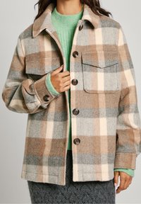 Mujer con una chaqueta de cuadros en tonos beige, gris y crema, sobre un suéter de punto verde y una falda oscura texturizada, sosteniendo el cuello de la chaqueta con la mano.