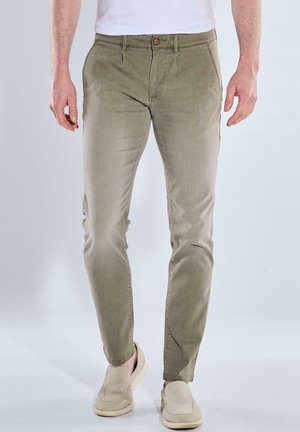 Mann trägt schlanke beigefarbene Hose mit Knopf- und Reißverschluss, kombiniert mit hellbeigen Slip-On-Schuhen, stehend vor einfachem Hintergrund.