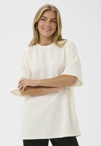 Vörös hajú nő mosolygik, karjait keresztbe tették, nagyméretű, fehér, rövid ujjú ingben és fekete nadrágban áll egy sima háttér előtt.