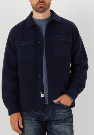 Giacca di lana blu navy con chiusura a bottoni, due tasche sul petto e una finitura testurizzata, abbinata a un maglione girocollo azzurro chiaro.