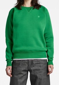 Sudadera verde con una textura suave, cuello redondo y puños acanalados. Marca mínima en la esquina superior izquierda. Combinada con pantalones gris oscuro.