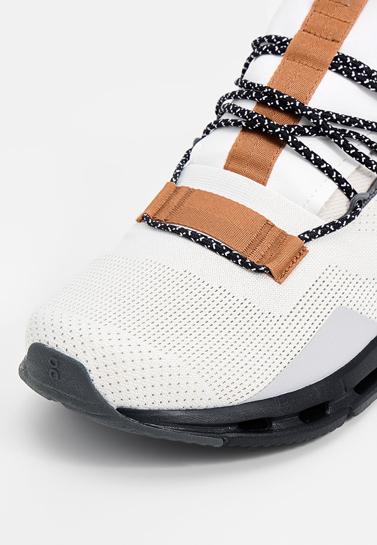 Weißer Sportschuh mit schwarz gesprenkelten Schnürsenkeln, braunen Stoffschlaufen, atmungsaktivem Mesh-Oberteil und schwarzer gepolsterter Sohle.