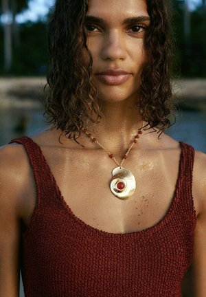 Donna con capelli ricci e bagnati indossa una canottiera rossa testurizzata e una collana d'oro con un grande pendente rotondo in pietra rossa all'aperto.