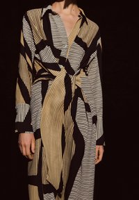 Person in einem langärmligen, geknöpften Kleid mit abstrakten schwarz-beigen Streifenmustern, das an der Taille zu einem Knoten gebunden ist, vor einem dunklen Hintergrund.