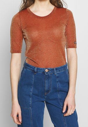 Femme portant un haut court pailleté couleur rouille, rentré dans un jean bleu taille haute, sur un fond uni.