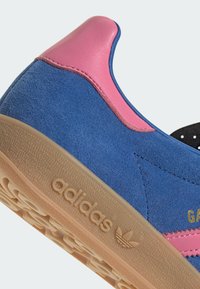 Modré semišové tenisky s ružovými koženými akcentmi, gumovou podrážkou a zlatým brandingom. Obsahujú textúrovaný zvršok a nízky profil.