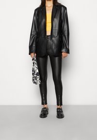 Černá kožená blazerka přes žlutý crop top, v kombinaci s černými koženými kalhotami se zipy na kotnících a černými loafers. Drží vzorovanou tašku.