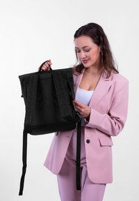 Černý batoh obdélníkového tvaru, s dvojitými rukojeťmi, několika popruhy a texturovanou zadní částí. Stylizováno proti jednobarevnému pozadí.