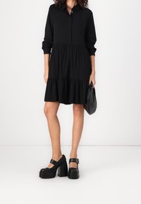 Abito nero con bottoni, orlo a strati, maniche lunghe e vestibilità rilassata. Abbinato a scarpe nere con plateau spesso e una piccola borsa nera.