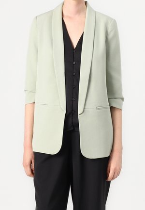 Personne portant un blazer vert clair aux manches retroussées sur une chemise noire boutonnée et un pantalon noir sur fond blanc.