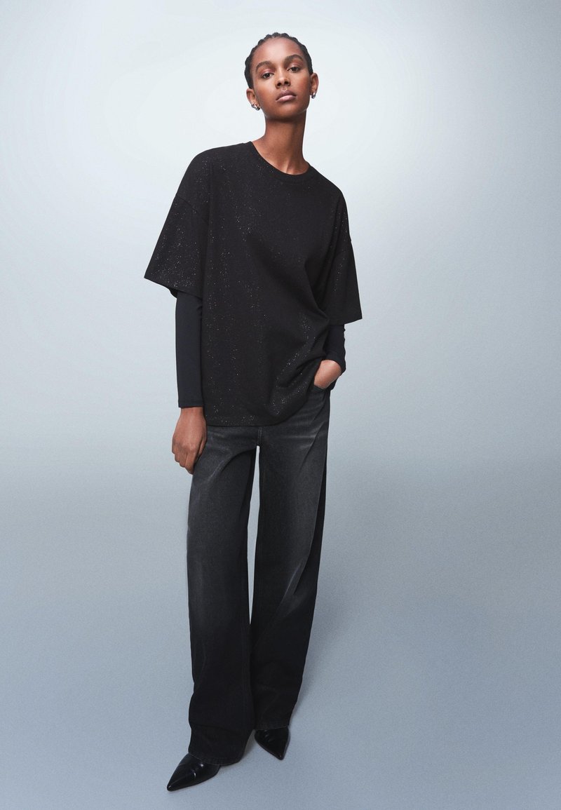 Black oversized t-shirt with subtle sparkles, paired with dark wide-leg jeans and pointed black shoes, set against a light background.
