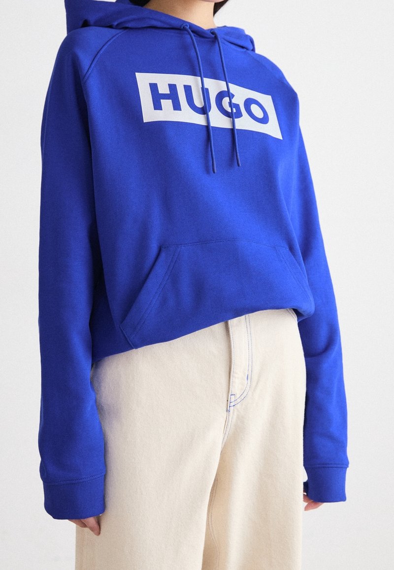 Personne portant un sweat à capuche bleu vif avec l'inscription « HUGO » sur la poitrine et un pantalon taille haute écru sur un fond uni.