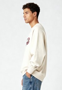 Sweat-shirt crème avec col rond, coupe ample et épaules tombantes ; présente un texte graphique rouge et noir à l'avant. Associé à un jean bleu.