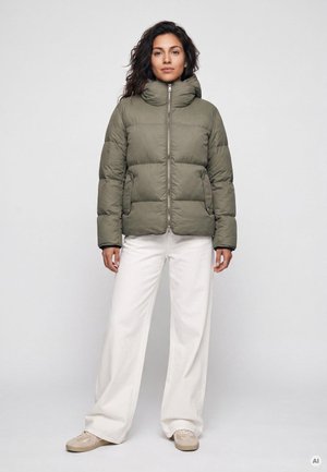 Frau mit dunklen Haaren, die eine grüne Daunenjacke, weiße weite Hose und beige Sneaker trägt, steht vor einem schlichten hellen Hintergrund.