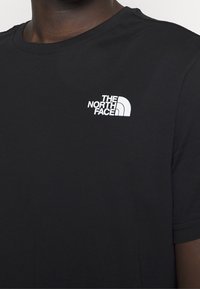 Camiseta negra de cuello redondo con el logotipo blanco de The North Face en la parte superior izquierda del pecho, llevada por una persona con tono de piel oscura.