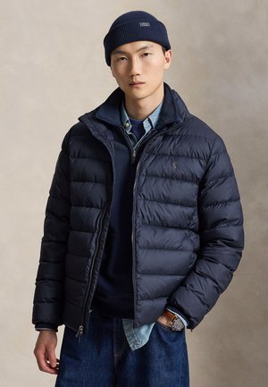 Homme portant un bonnet bleu marine, une doudoune bleu marine, une chemise en denim, un pull, un jean et une montre-bracelet, debout devant un fond beige uni.