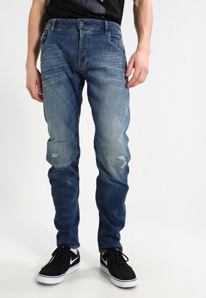 Mężczyzna ubrany w wyblakłe, postarzane niebieskie dżinsy i czarne buty sportowe Nike, stojący na tle gładkiego białego tła.