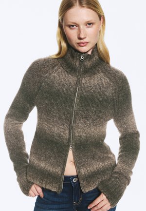 Femme portant un pull à fermeture éclair marron dégradé et duveteux avec un col montant et un jean bleu, debout les mains sur les hanches sur fond blanc.