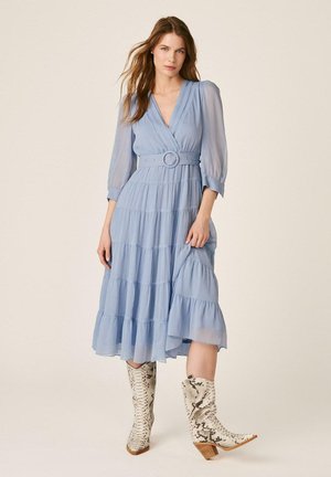 Mujer con vestido midi azul claro de capas, con un cinturón ancho y mangas transparentes, combinado con botas blancas hasta media pantorrilla con estampado de piel de serpiente.