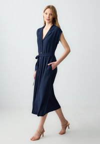Granatowa sukienka midi z krótkimi rękawami, dekoltem w kształcie litery V, paskiem w talii i bocznymi kieszeniami; wykonana z lekkiego materiału; z eleganckimi detalami w postaci guzików.