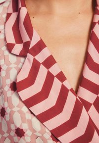 Apykaklė su geometriškais raštais rožinės ir bordo spalvų atspalviuose, pagaminta iš lygios medžiagos ir su ryškiomis chevrono detalėmis.