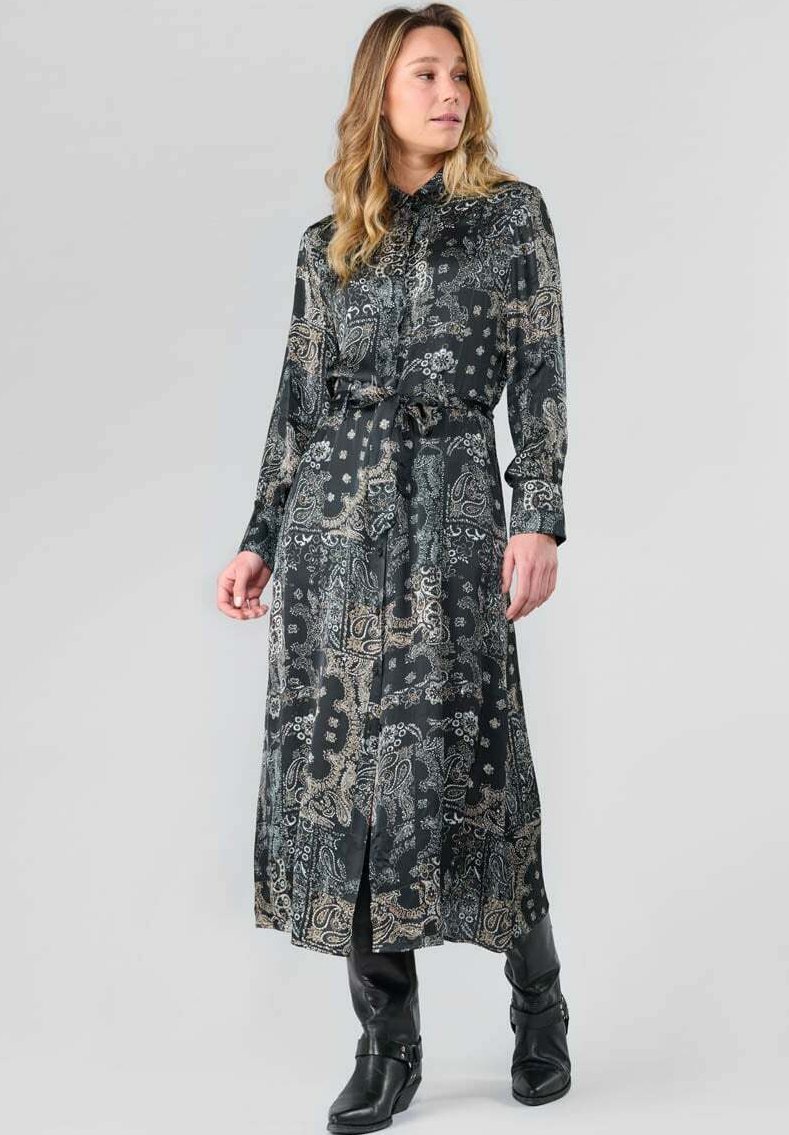 Lang zwarte jurk met paisley- en bloemenpatronen in goud en crème. Heeft een knoopsluiting aan de voorkant, een ceintuur in de taille en lange mouwen. G gedragen met zwarte laarzen.