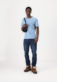 Light blue t-shirt with short sleeves and a small logo, paired with dark blue jeans and brown shoes with beige accents, holding a black bag.