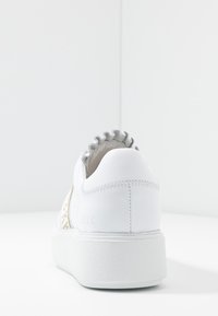 Bílé tenisky s texturovanou podrážkou, volánkovým límcem kolem kotníku a jemným prošíváním, zobrazené zezadu na hladkém bílém pozadí.