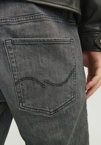 Des jeans en denim gris présentent une poche arrière avec un détail de couture noire incurvée. Le tissu a une texture douce et une apparence délavée.