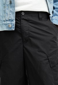 Shorts cargo noirs en tissu léger avec une texture lisse, dotés de poches latérales et d'un léger éclat. Associés à une veste en denim.