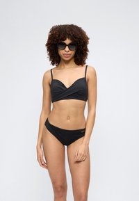 Γυναίκα με σγουρά μαλλιά, που φοράει μαύρο μπικίνι και μεγάλα μαύρα γυαλιά ηλίου, στέκεται μπροστά σε απλό φωτεινό φόντο.