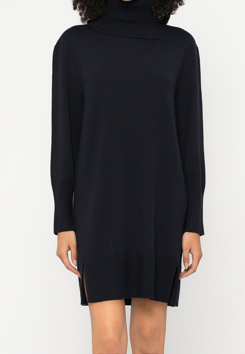 Femme portant une robe-pull noire à manches longues avec col roulé, avec des poignets et un ourlet côtelés, et des fentes latérales au-dessus des genoux.