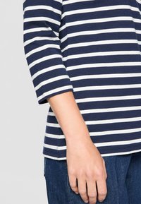 Tamsiai mėlynos ir baltos juostelės, medvilnės mišinio marškiniai su trimis ketvirtadaliais rankovėmis, apvalia iškirpte ir laisvu siluetu. Rankos poilsis ant tamsaus džinsinio audinio.