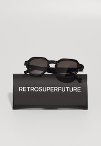 Occhiali da sole neri con montatura ottagonale, lenti scure e finitura lucida, appoggiati su un astuccio nero con la scritta bianca "RETROSUPERFUTURE."