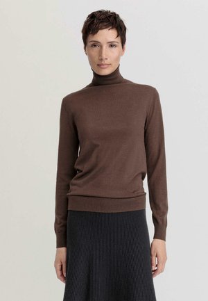 Maglione lungo a maniche con colletto alto di colore marrone, realizzato in un materiale morbido, con un design aderente e orlo a coste. Indossato con una gonna scura e strutturata.